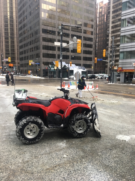 ATV equipped for snow removal, parked and ready to begin clearing snow in front of a commercial building